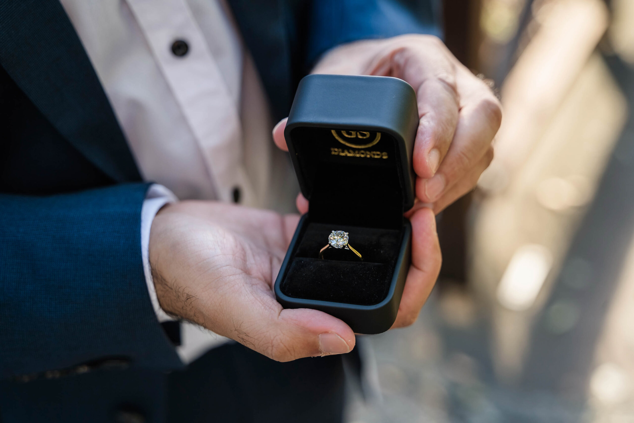 surprise engagement proposal, with gentleman in a suit holding the diamond engagement ring that he is about to propose with.